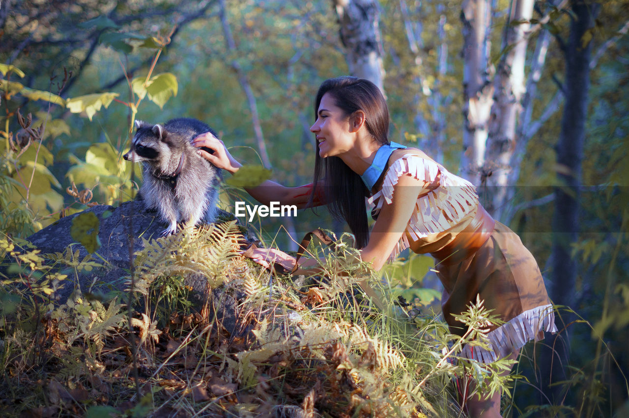 Side view of young woman in traditional clothing stroking raccoon in forest