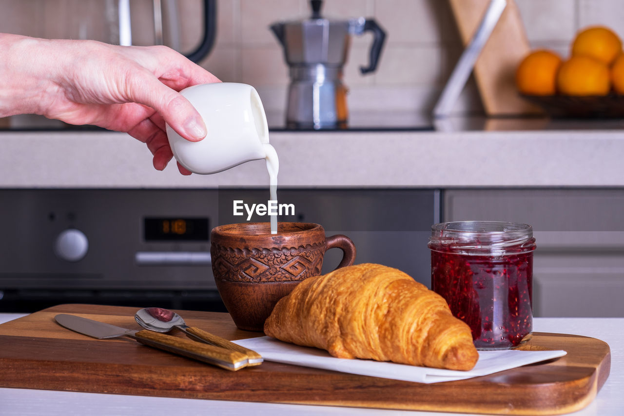 Served morning breakfast with croissant, jam and cup of coffee pouring cream in woman's hand