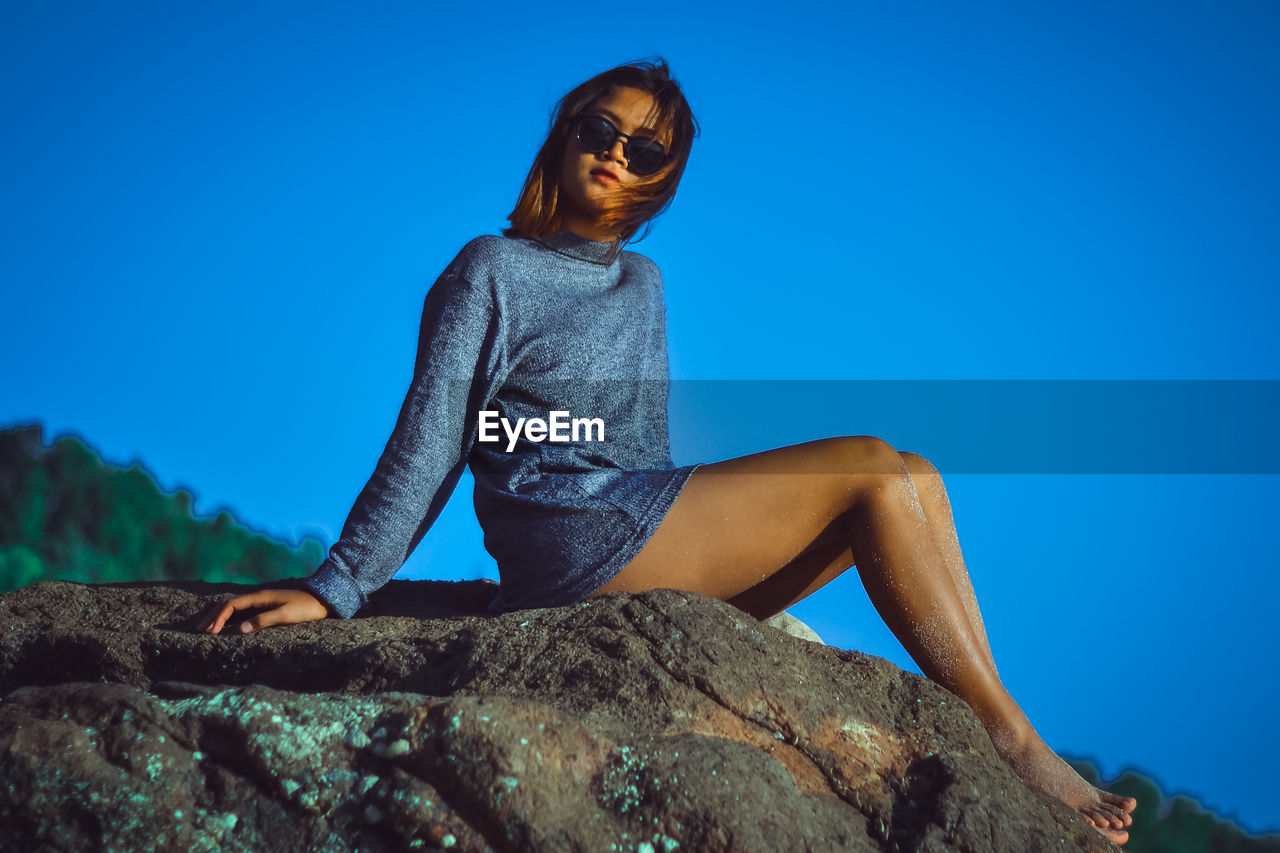 Low angle view of woman sitting on rock against clear blue sky