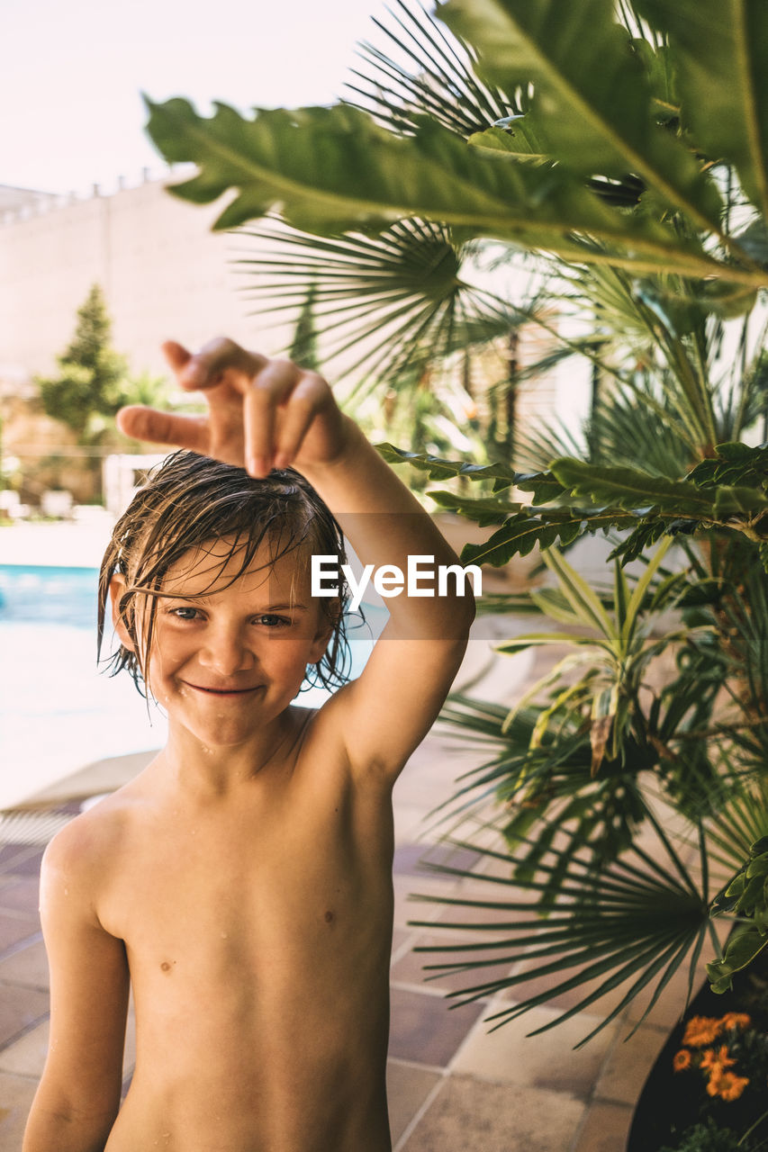 Portrait of shirtless boy standing by plant against swimming pool in resort