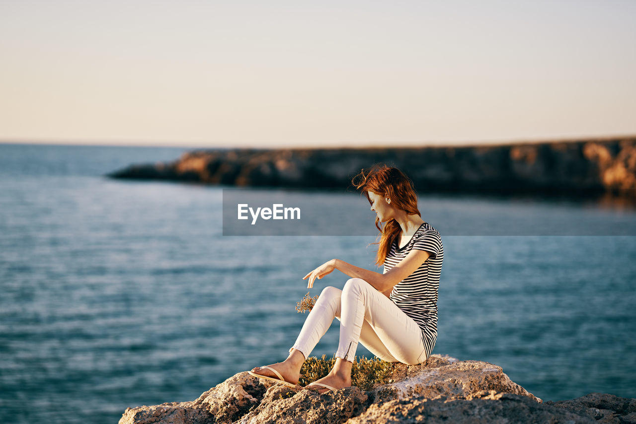 WOMAN SITTING ON ROCK LOOKING AT SEA SHORE AGAINST SKY