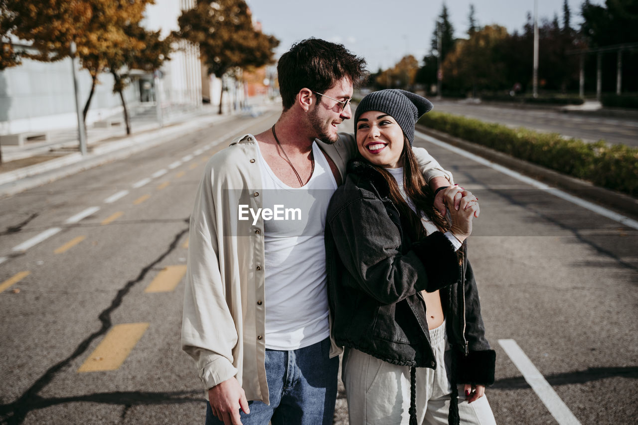 Smiling woman holding hands with male partner while standing on road in city