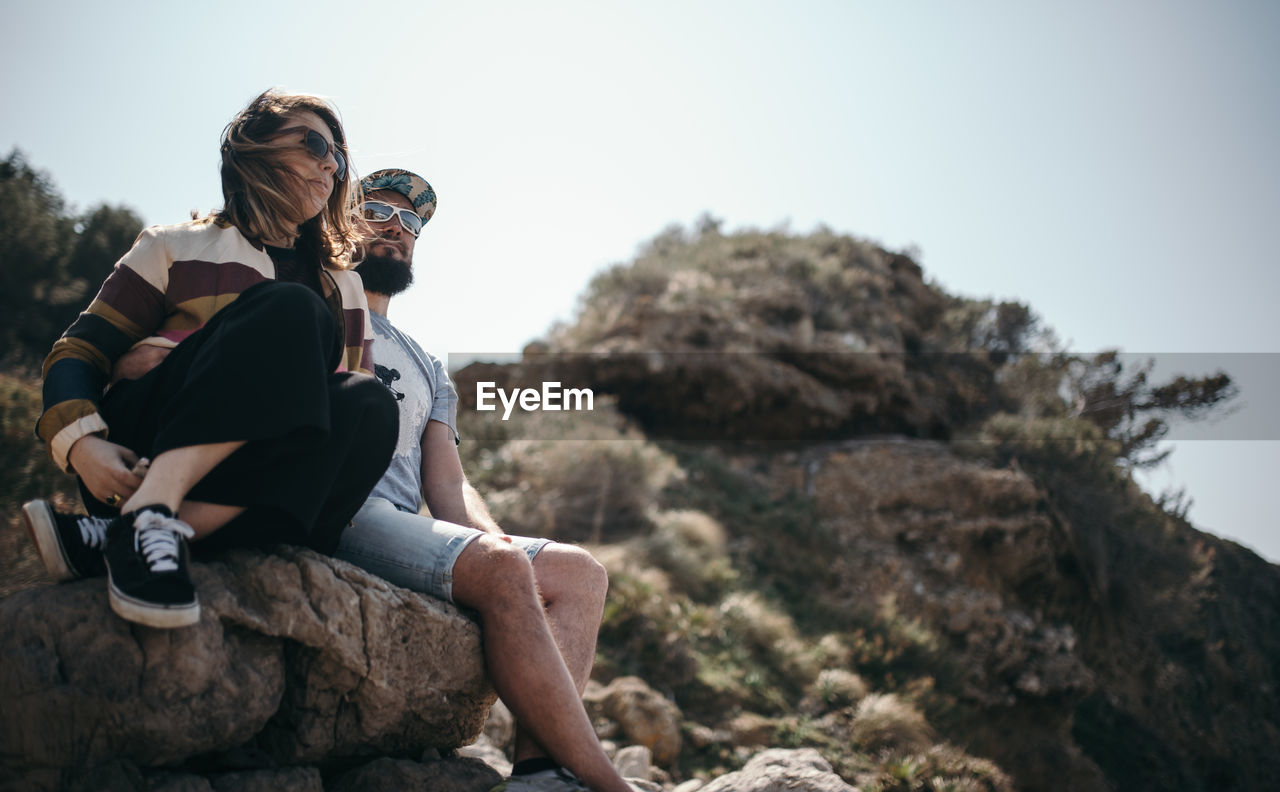 Low angle view of couple sitting on rock against clear sky