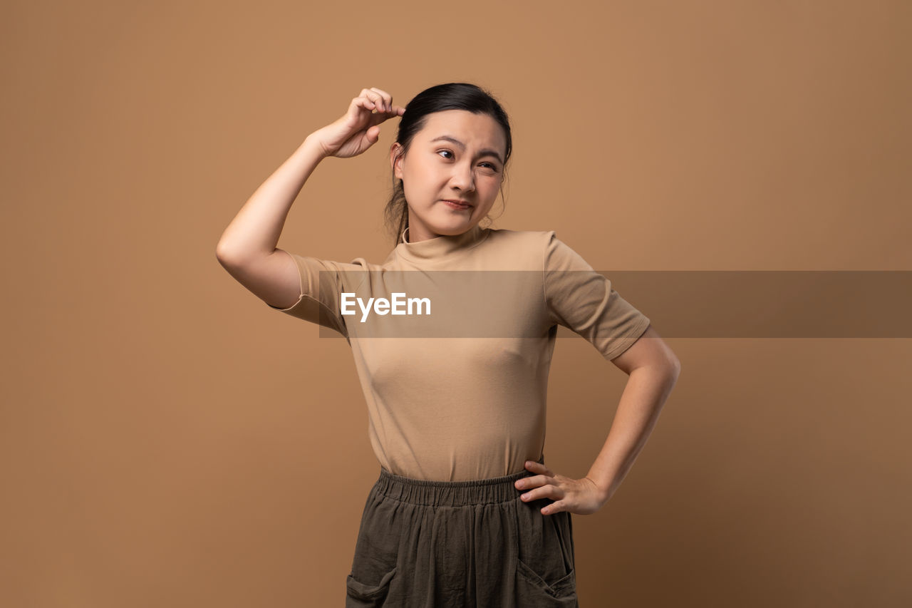 studio shot, one person, colored background, adult, portrait, indoors, photo shoot, standing, looking at camera, hand, young adult, front view, emotion, copy space, beige background, clothing, arm, waist up, women, three quarter length, brown hair, arms raised, looking, brown, hairstyle