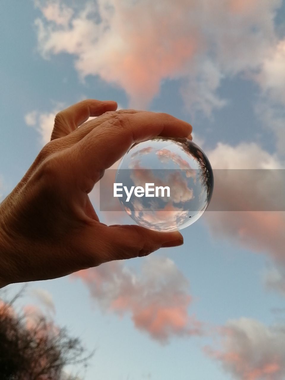 Low angle view of a hand holding up a crystal ball against a blue sky with pink clouds. 
