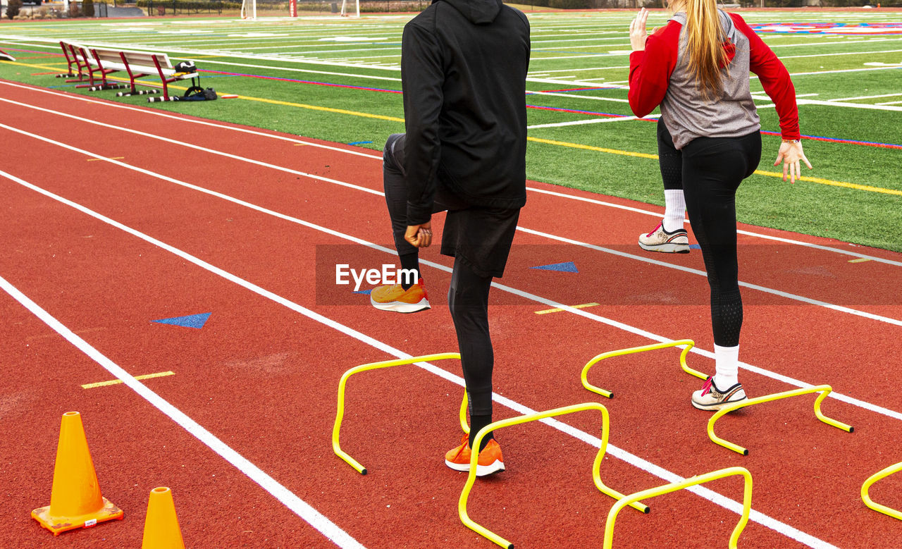 Rear view of two high school runners in a a-position over yellow mini hurdles on a red track.