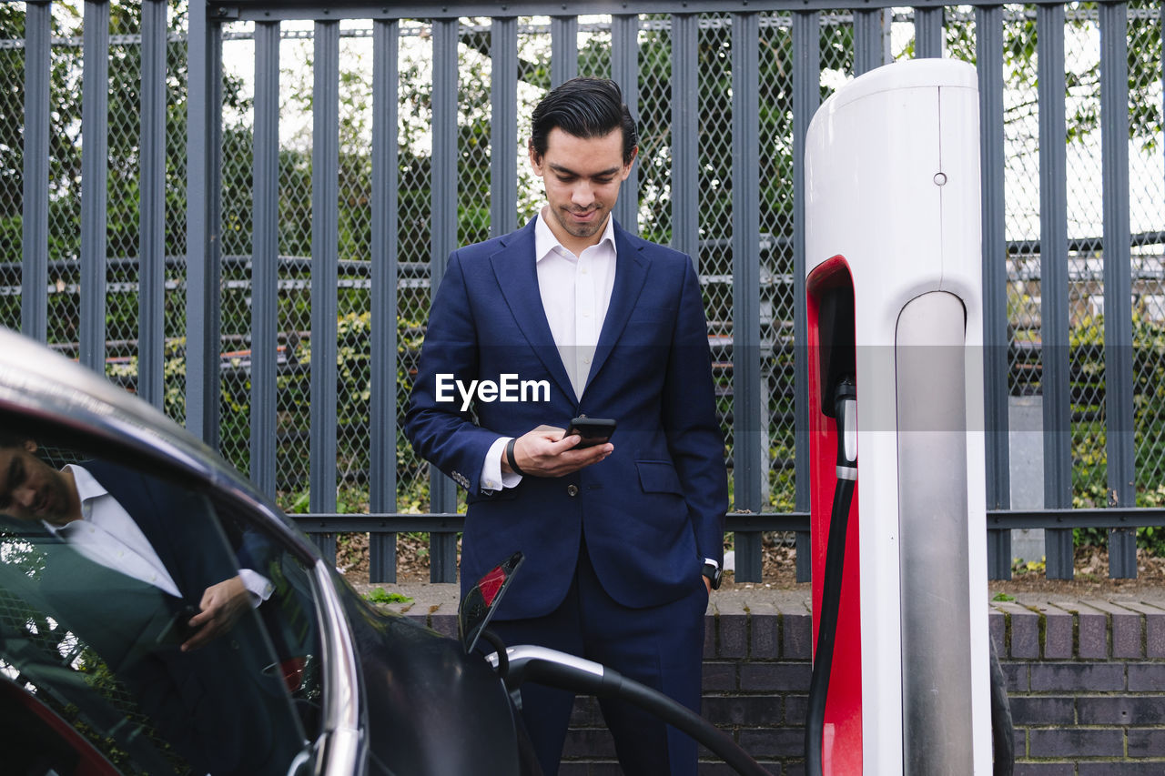 Businessman using smart phone standing at electric vehicle charging station