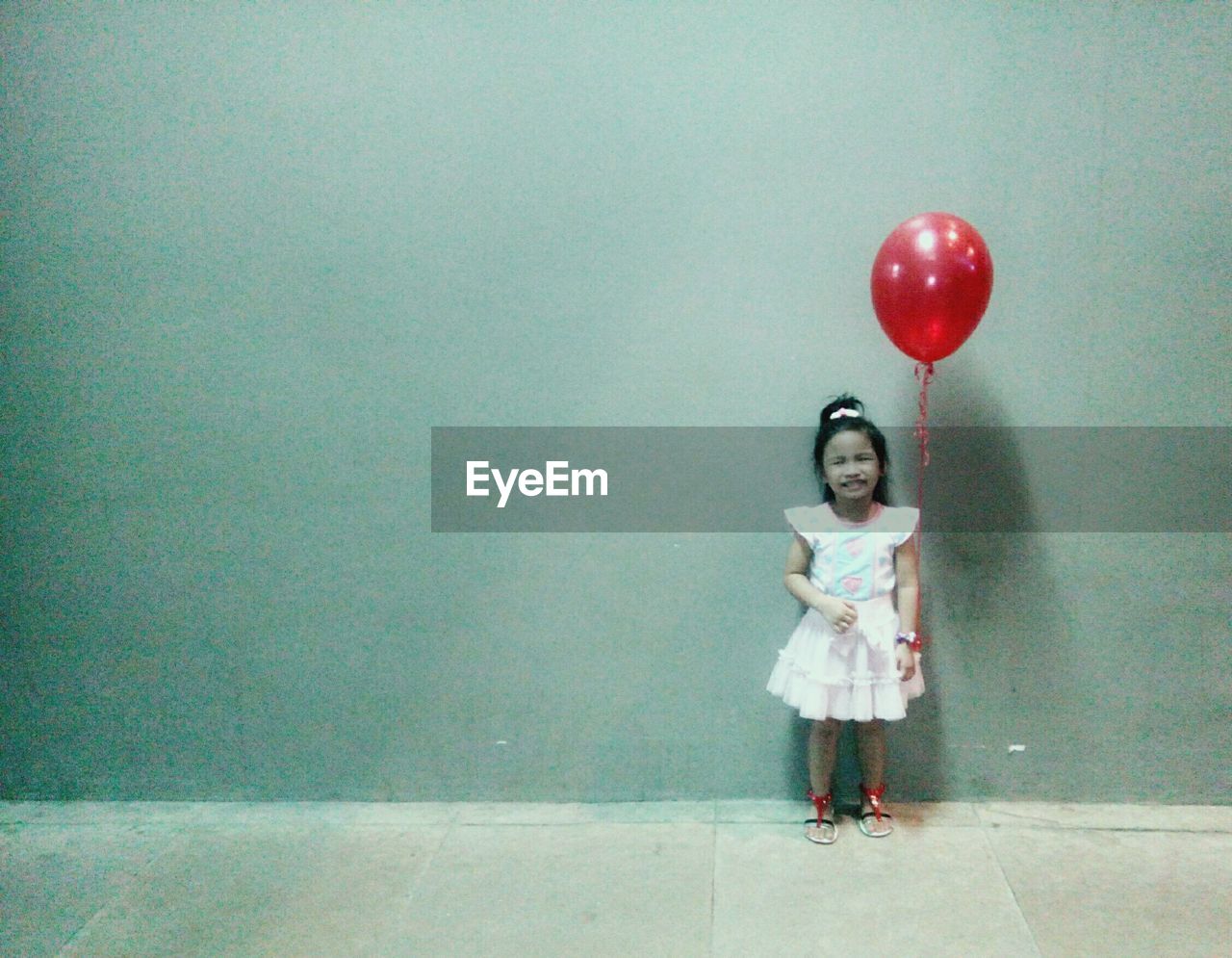 Happy girl with red balloon standing against wall