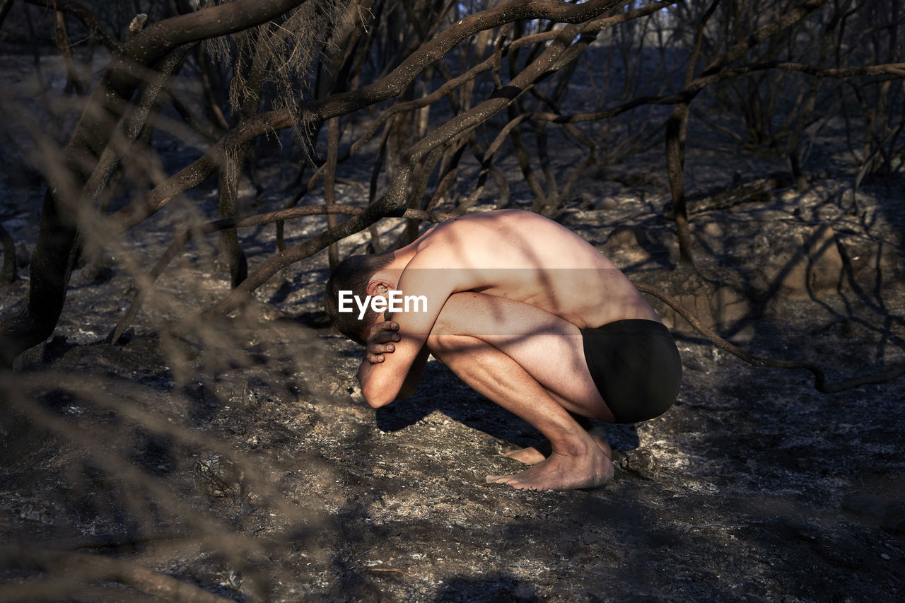 Shirtless man crouching on forest floor