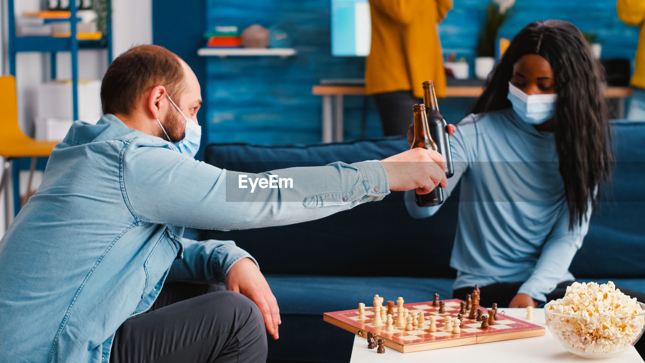 Couple wearing mask toasting beer while playing chess