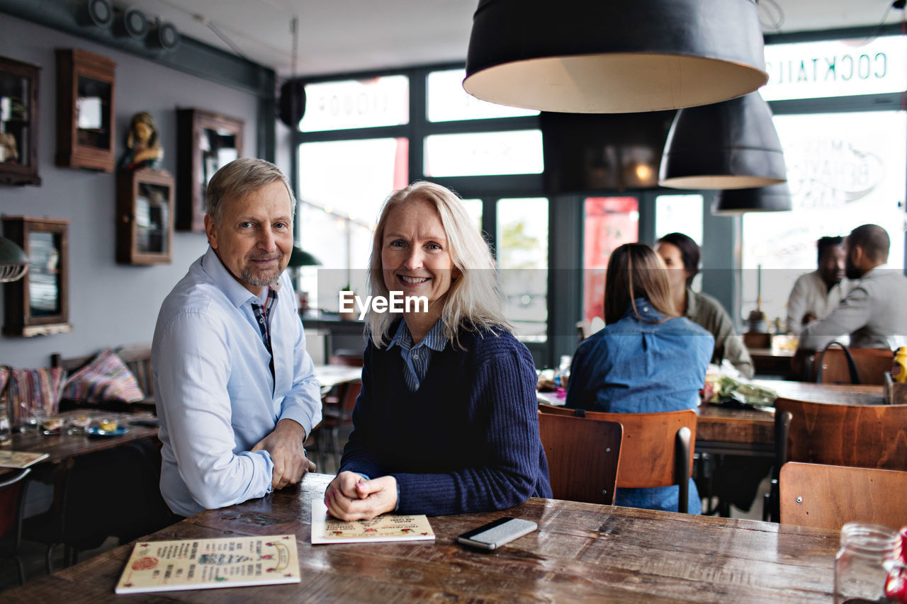 Portrait of smiling mature couple with menu at wooden table against people in restaurant