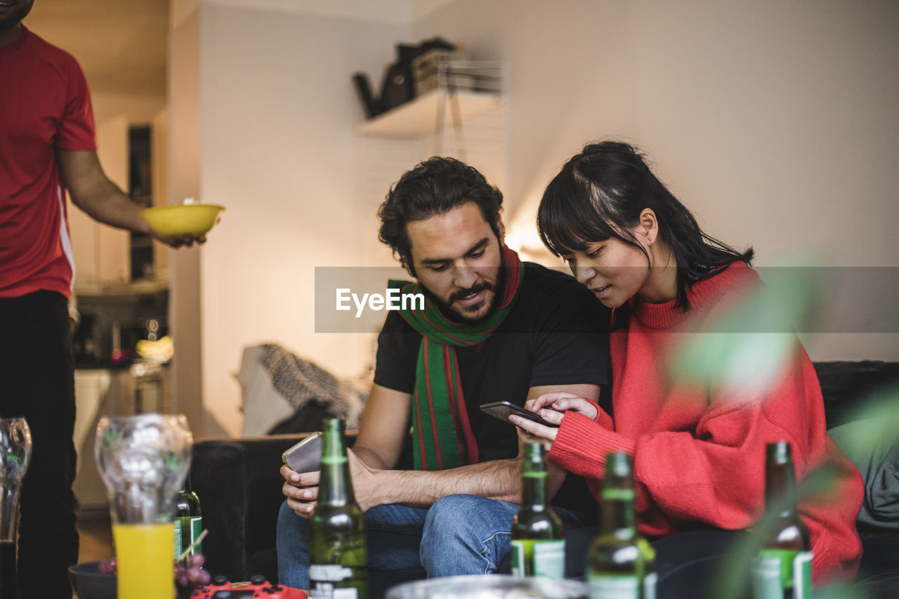 Male and female friends using smart phone while watching match at home