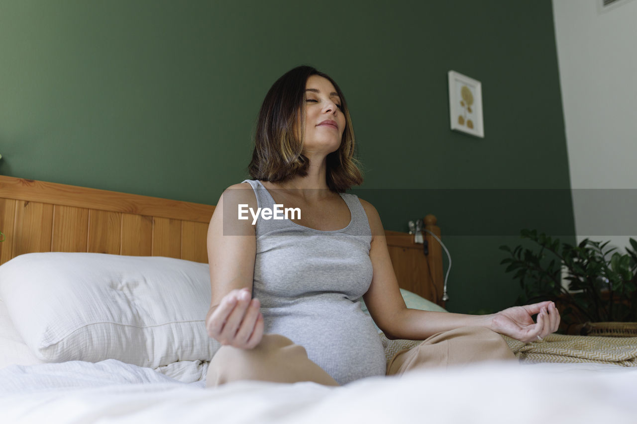 Pregnant woman with eyes closed meditating on bed at home
