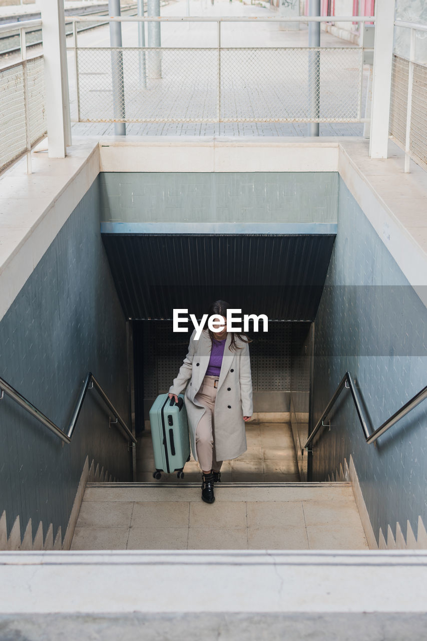 From above of elegant stylish young female traveler with suitcase walking up stairs at train station