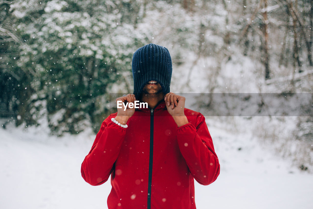 Smiling young man covering face with knit hat in forest during snowfall