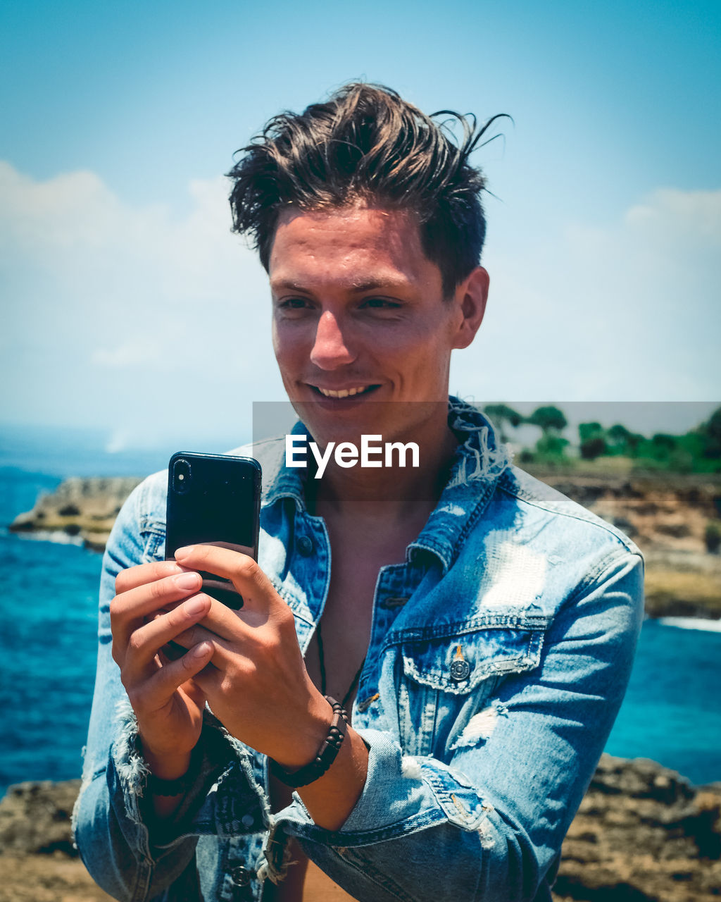Smiling man using smart phone at beach against sky