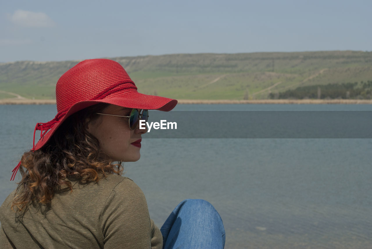 Woman in red cap and sunglasses sitting against lake