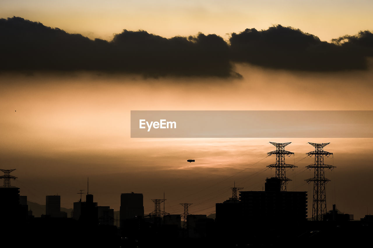 Cityscape with electricity pylons against cloudy sky during sunset