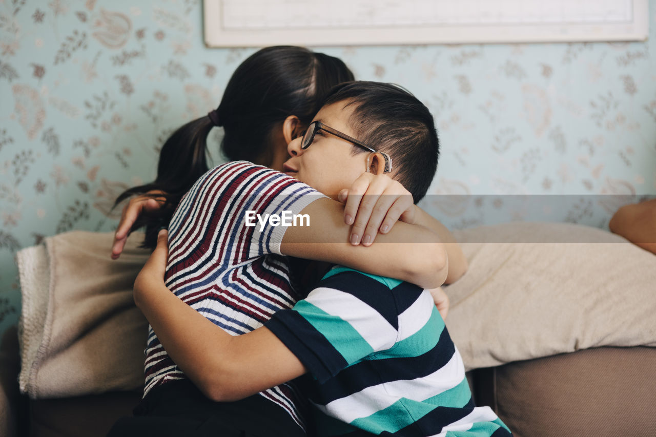 Sister embracing boy while sitting on sofa against wall at home