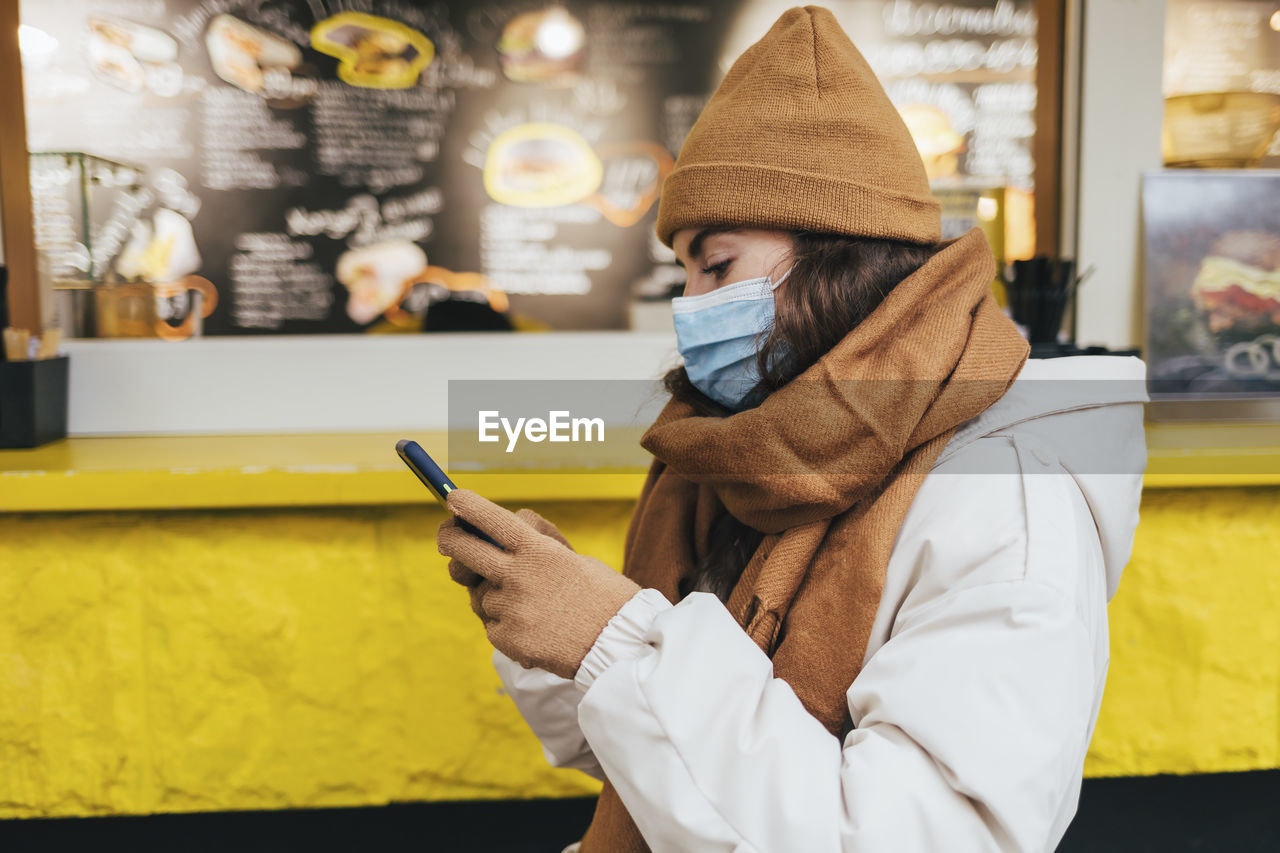 Young woman using smart phone waiting outside cafe in winter during covid-19