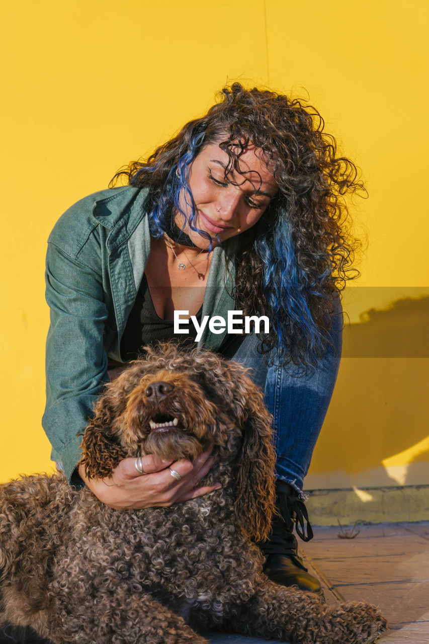 Woman with curly hair stroking water dog in front of yellow wall