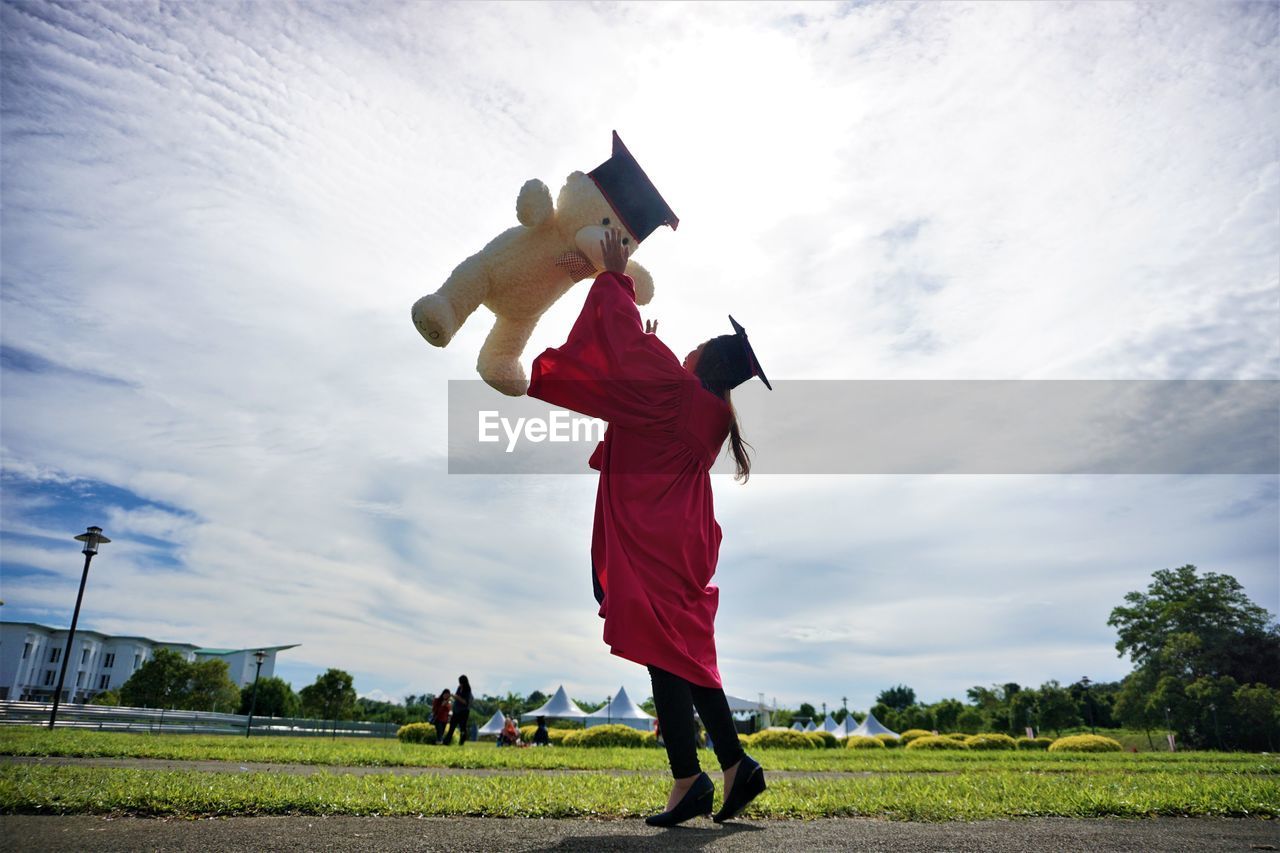 University student throwing teddy bear against sky during graduation