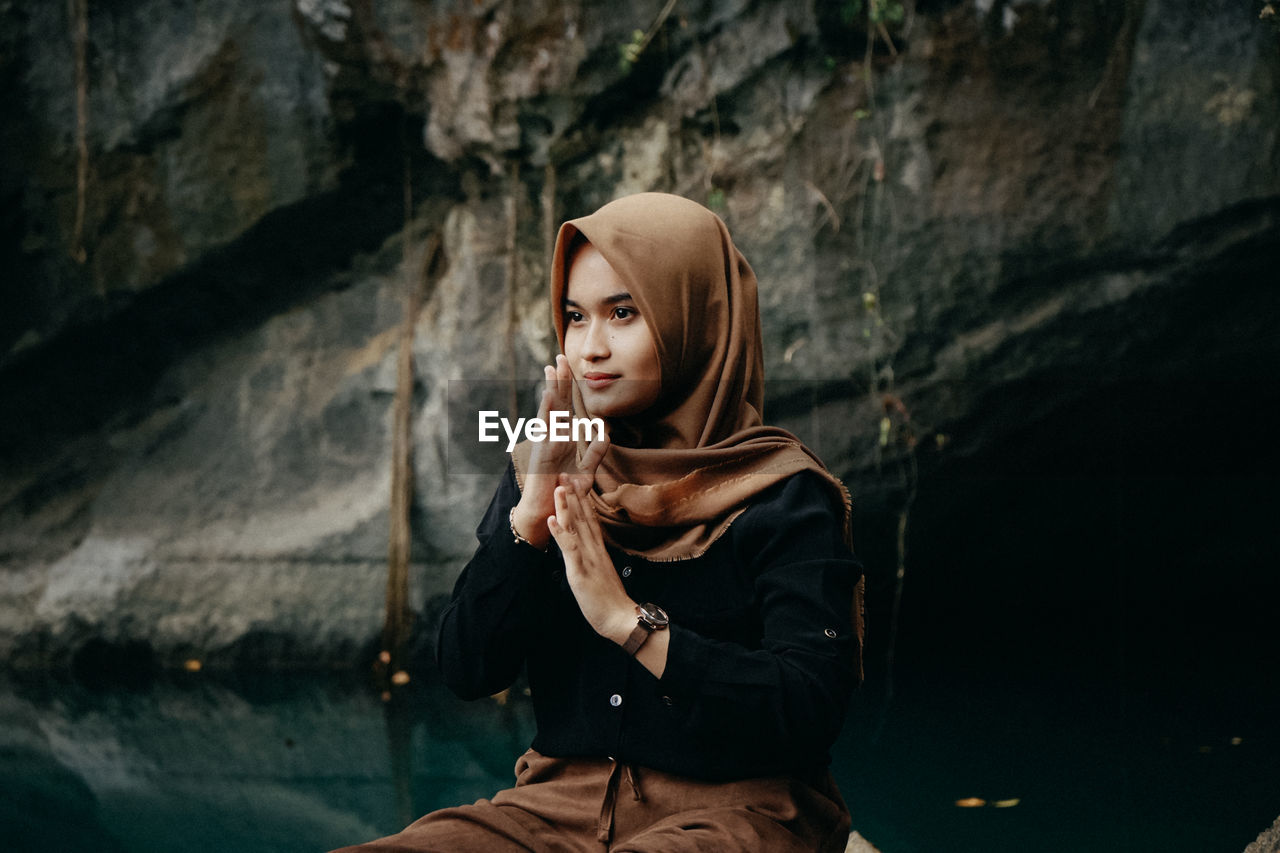 Young woman wearing hijab looking away while sitting against rock formation