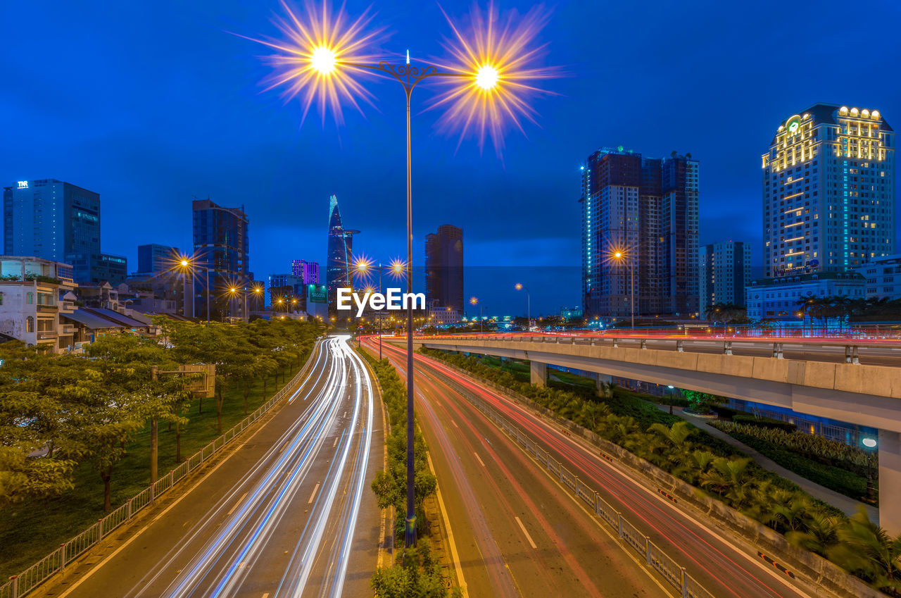 LIGHT TRAILS ON ROAD AMIDST ILLUMINATED BUILDINGS AGAINST SKY AT NIGHT