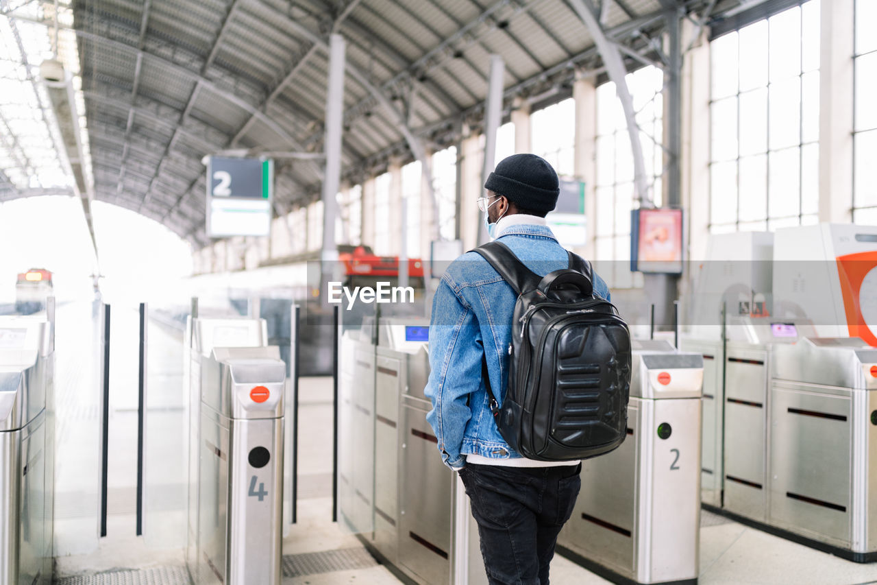 Back view of african american male tourist with backpack passing through turnstile on railway station while traveling during coronavirus pandemic