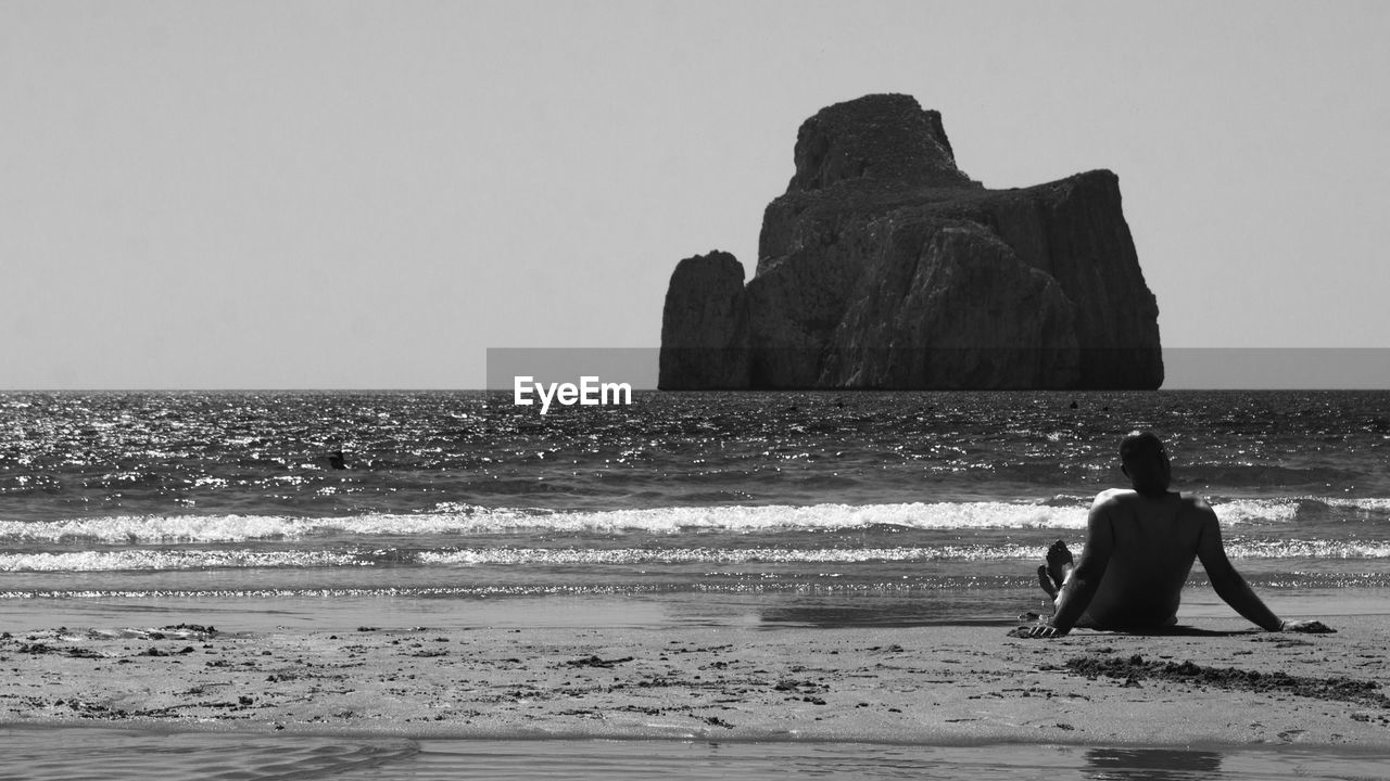 REAR VIEW OF MAN STANDING ON ROCK AT BEACH