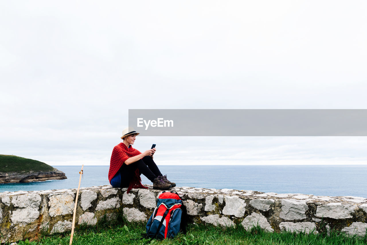 Full length side view of young female backpacker with trekking stick sitting on stone border near sea and browsing smartphone while resting and checking route during pilgrimage through spain