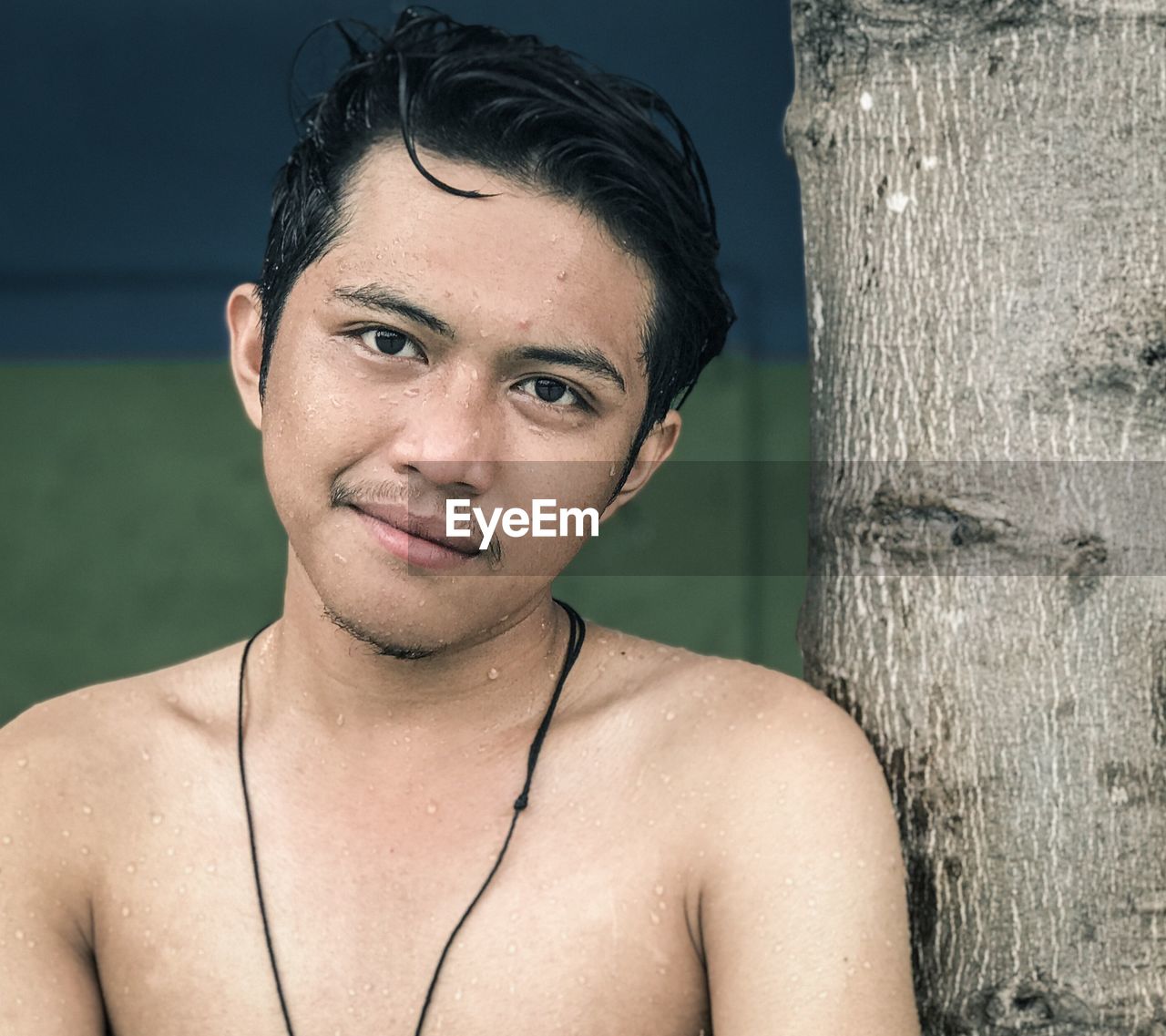 Portrait of shirtless man smiling while standing by tree trunk