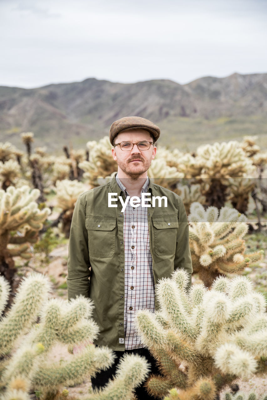 Young man with cap in cholla cactus field with hills in the background