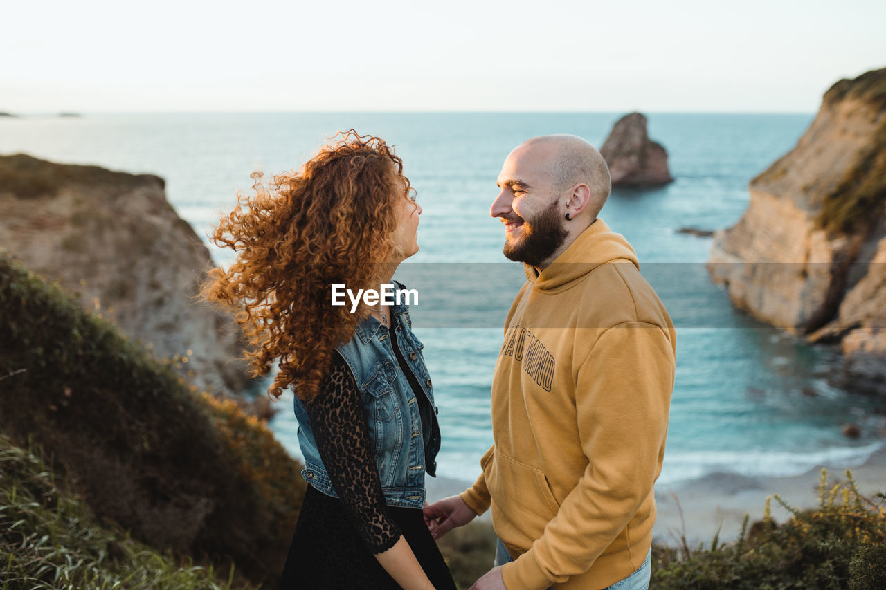 Side view of happy hipster couple holding hands and talking while standing on rocky seashore and spending sunny evening together