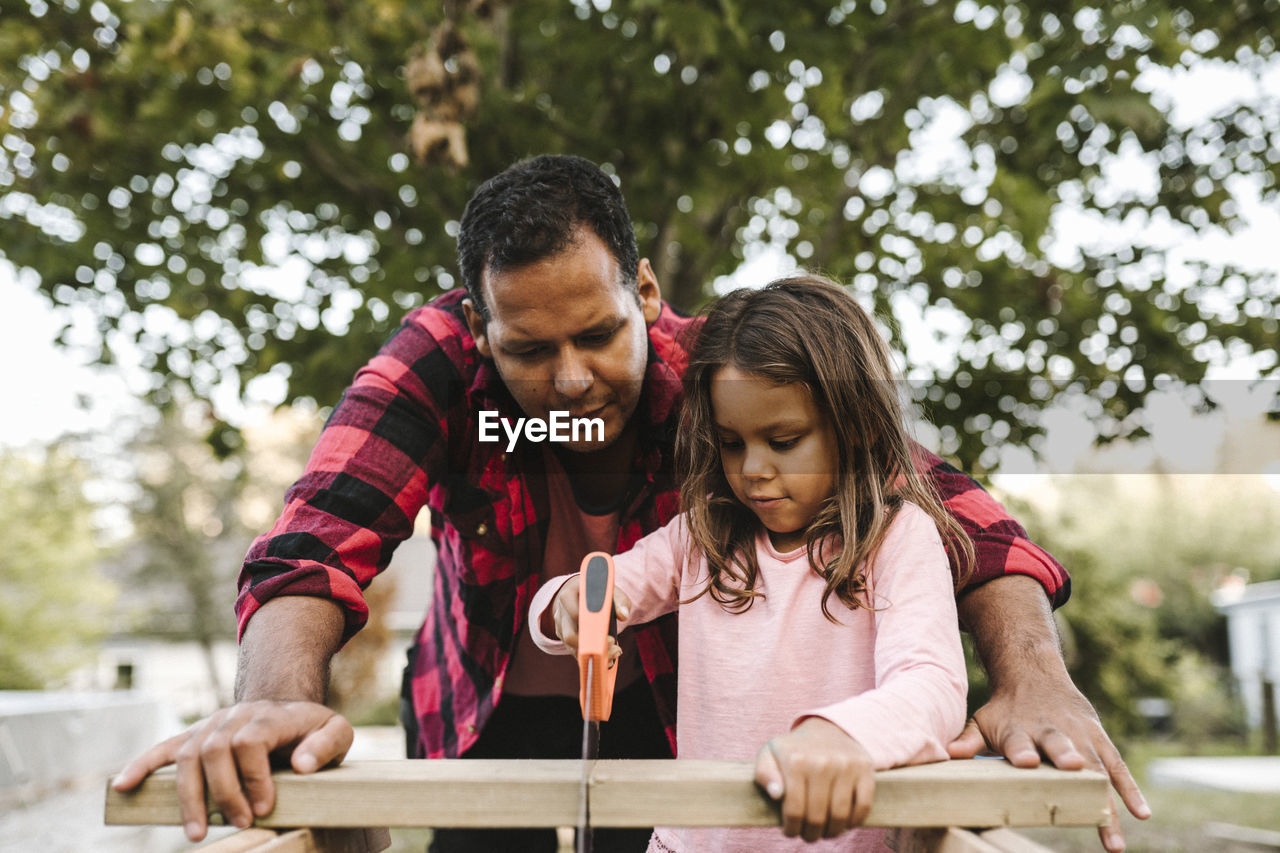 Girl cutting wooden plank by father during summer