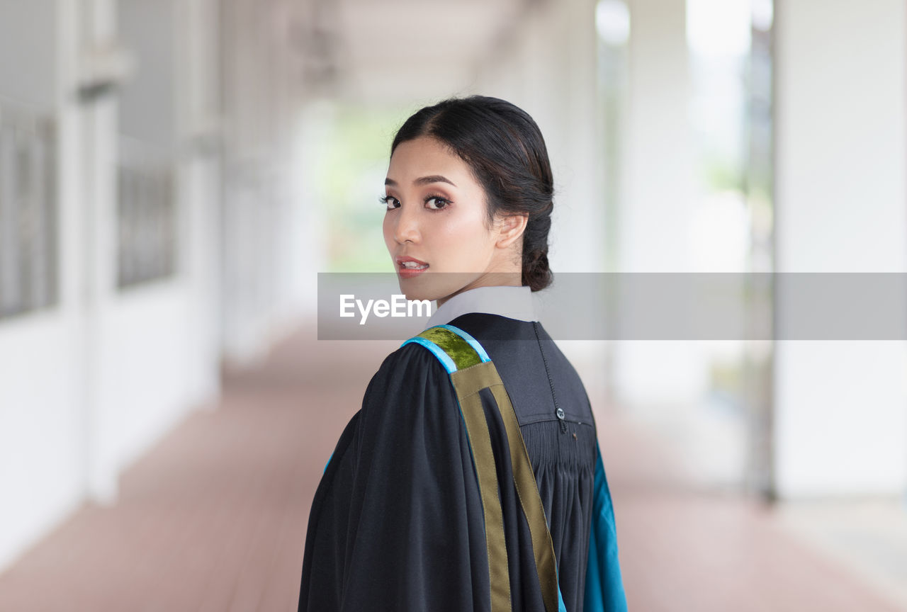 Portrait of young woman wearing graduation gown standing in corridor at university