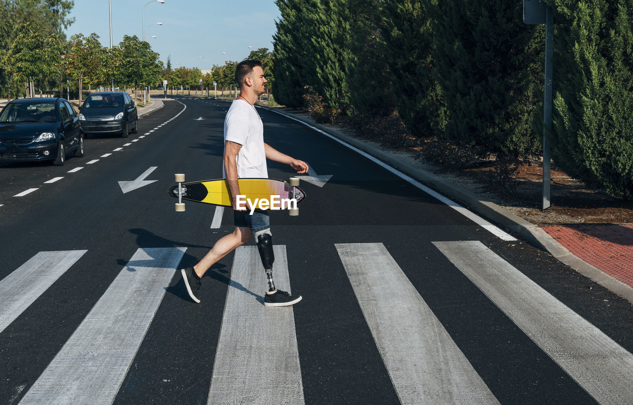 Young man with leg prosthesis holding skateboard and walking on crosswalk