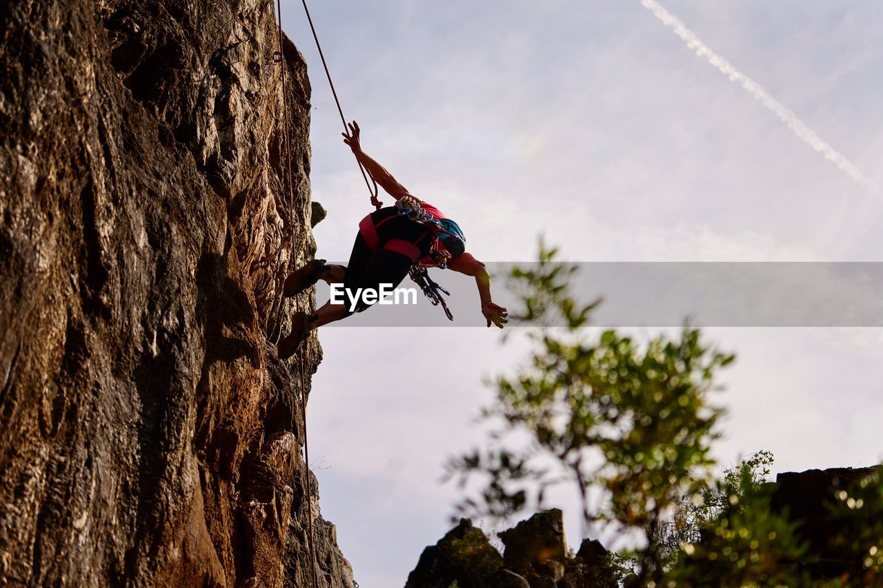 Low angle full length of active senior woman climbing on rocky cliff against sky