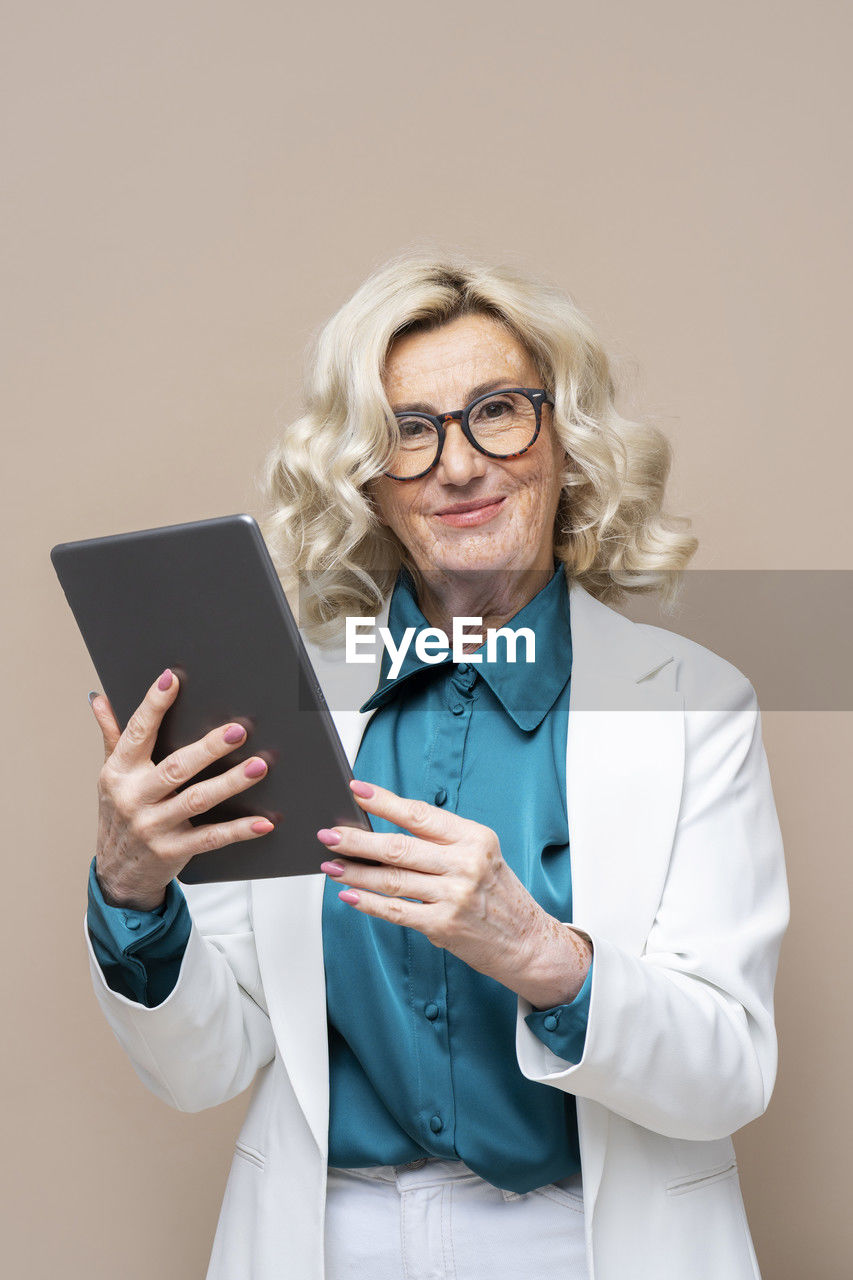 Smiling senior businesswoman holding tablet pc against beige background