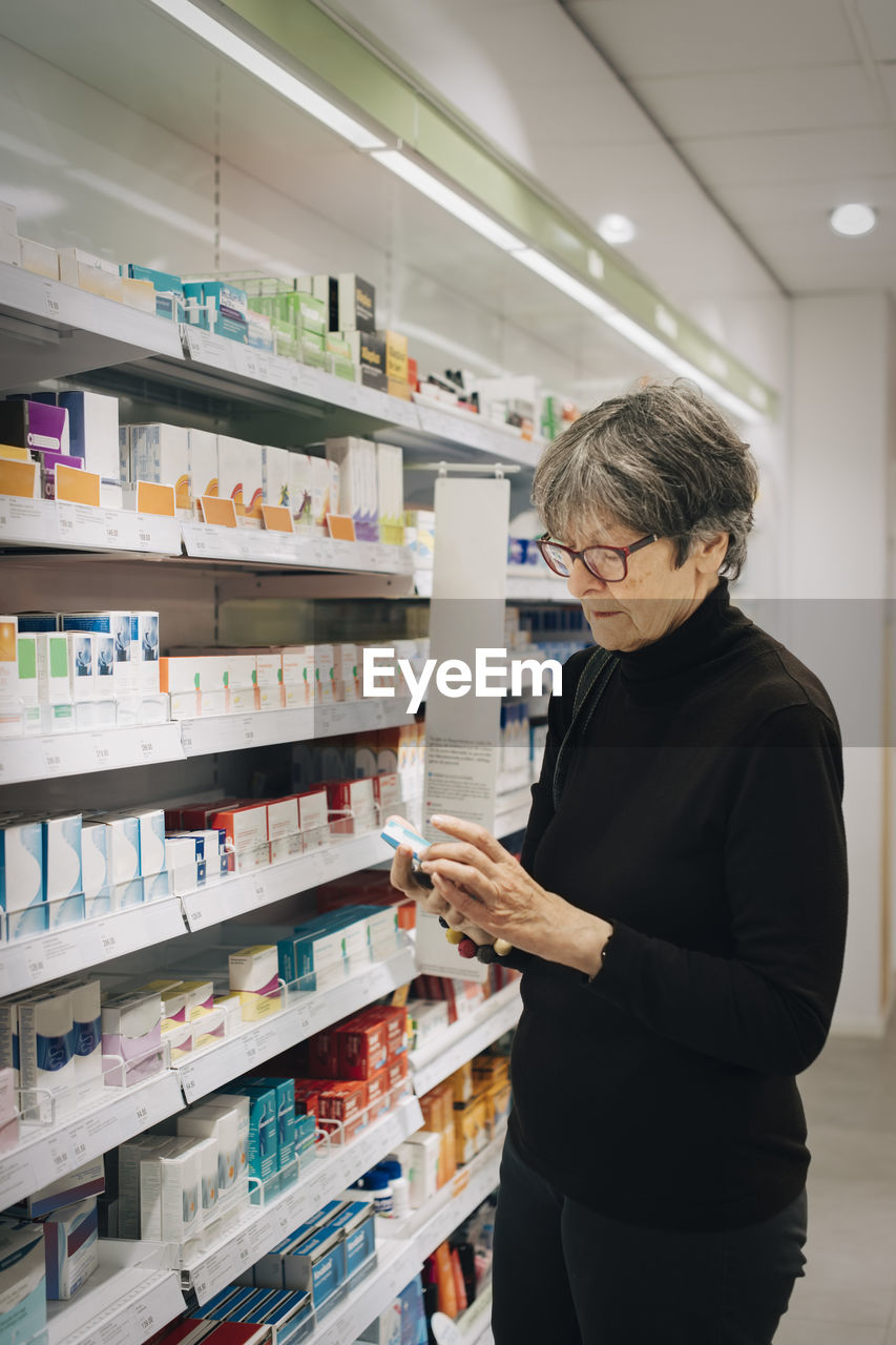 Senior female customer examining medicine by rack at pharmacy store