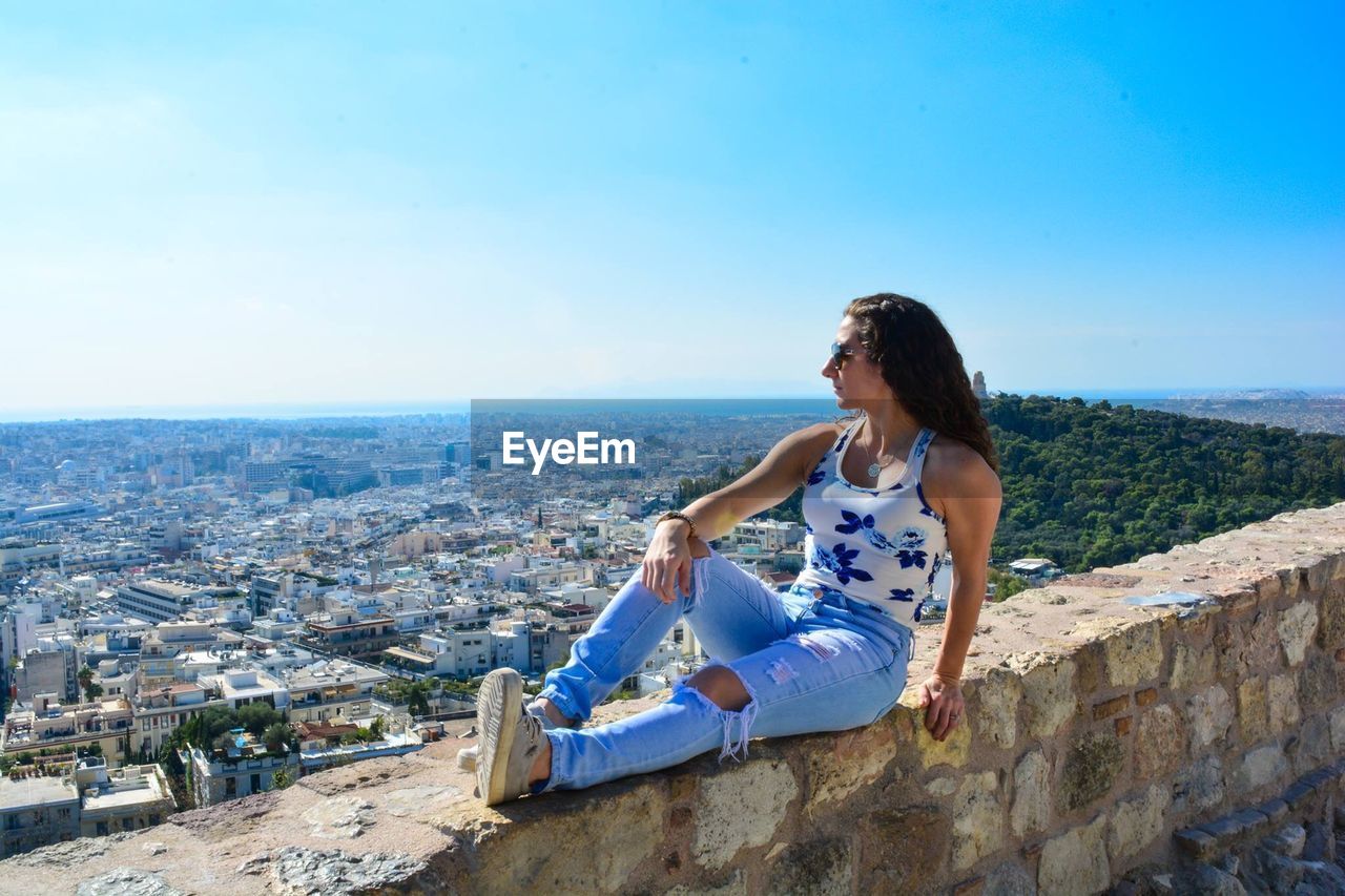 Young woman wearing sunglasses sitting on retaining wall against cityscape during sunny day