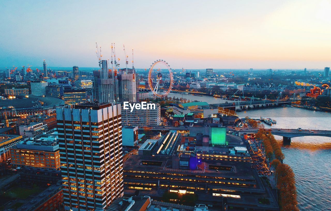 Aerial view of river amidst illuminated city against sky during sunset