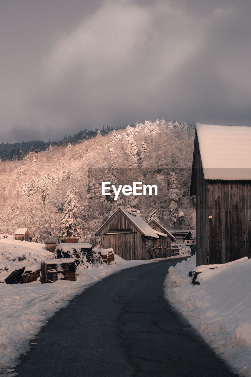 Road amidst wooden buildings against forest, mountain and sky during winter