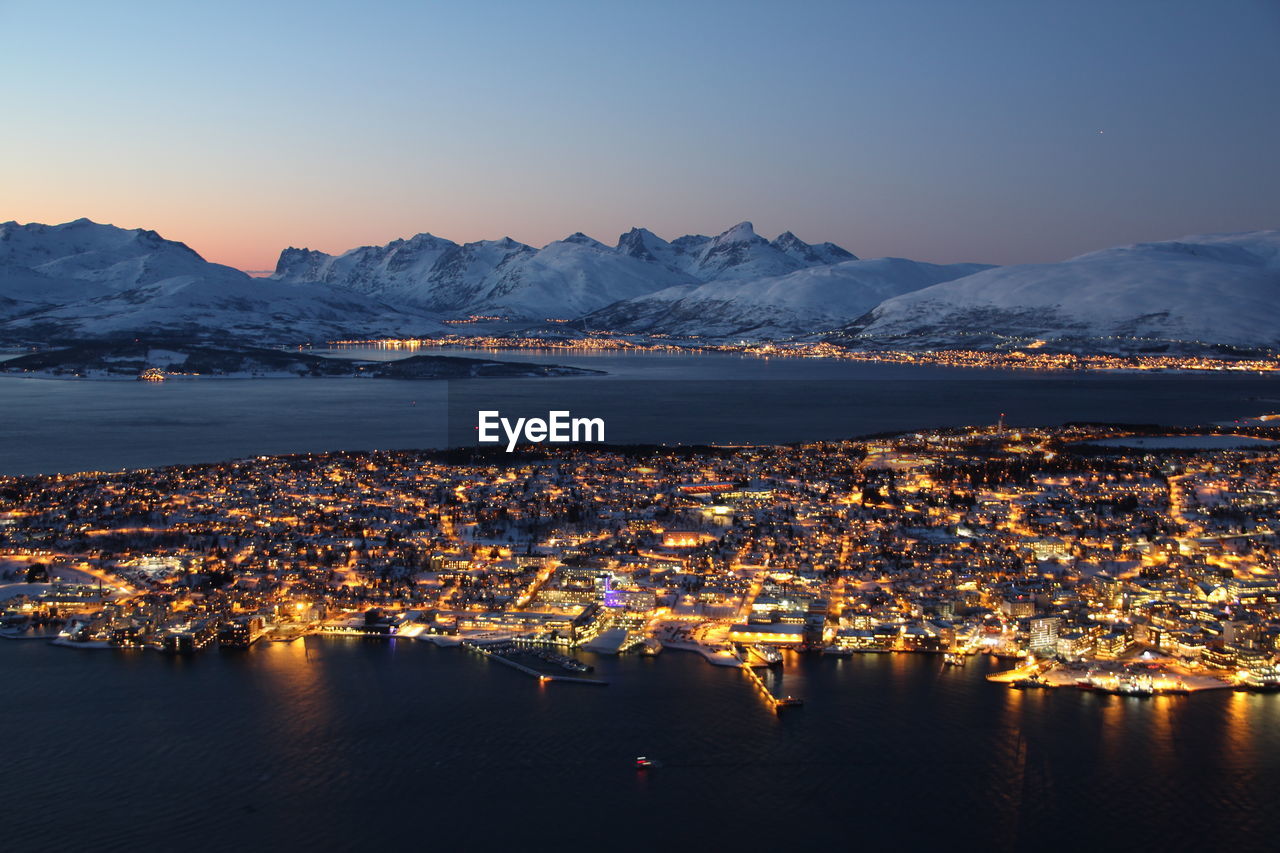 Illuminated cityscape by sea and snow covered mountains against clear sky at dusk