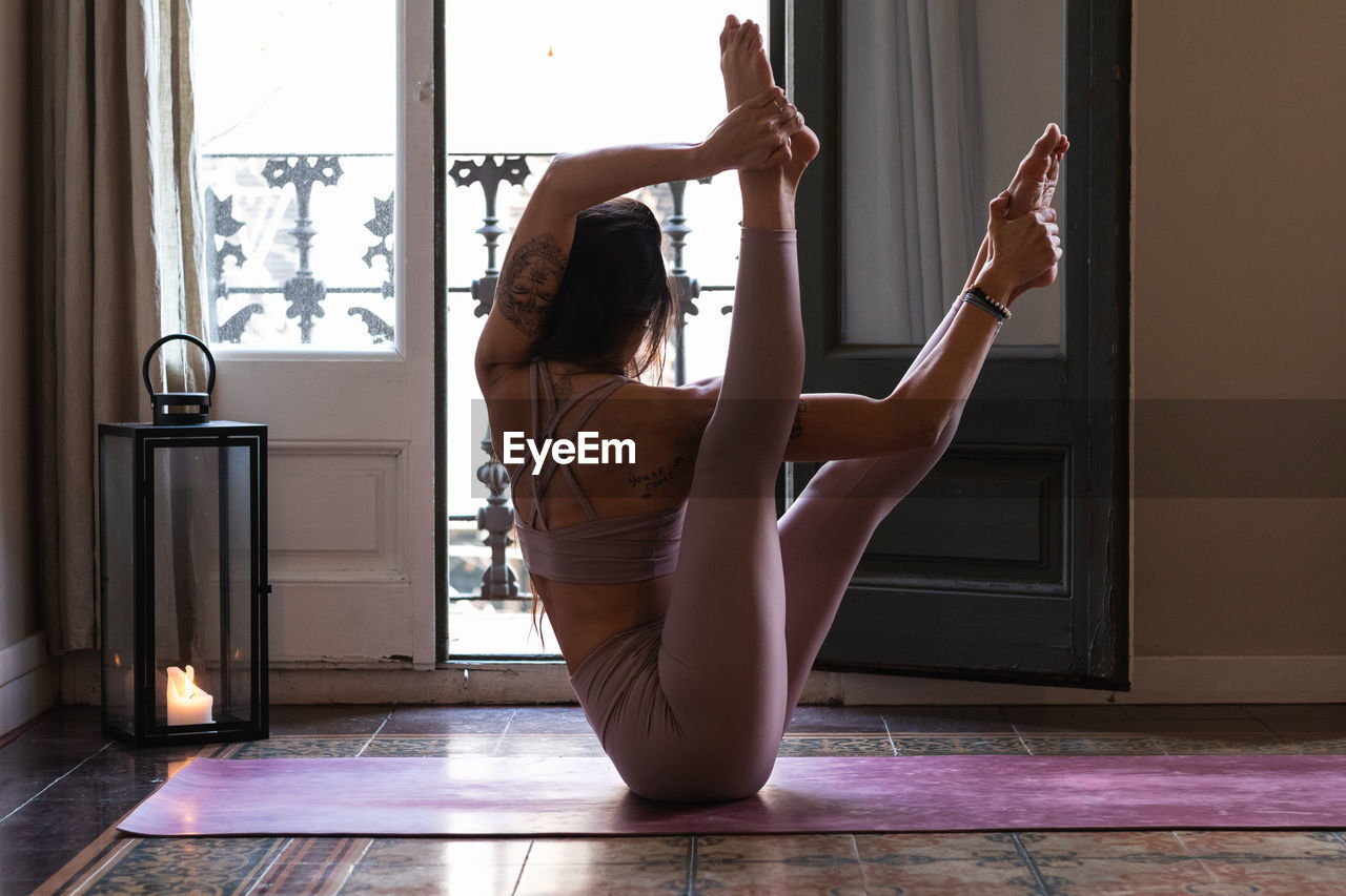 Side view of anonymous tattooed female practicing yoga with raised legs on mat in house