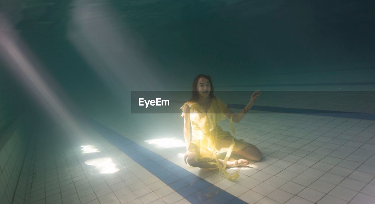Full length of young woman sitting on tiled floor underwater in swimming pool