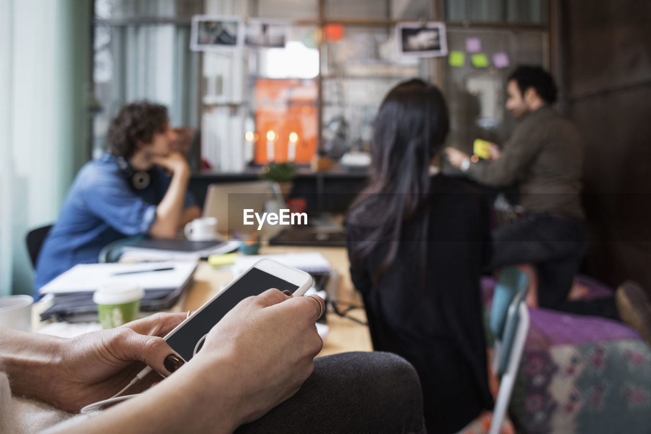 Close-up of woman holding mobile phone against colleagues working in creative office