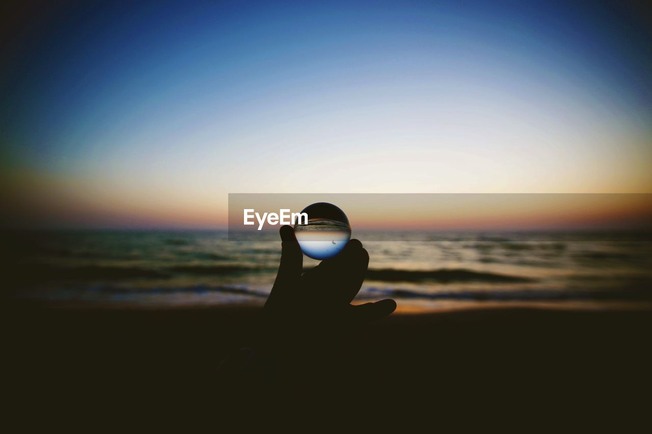 Cropped hand holding crystal ball at beach during sunset