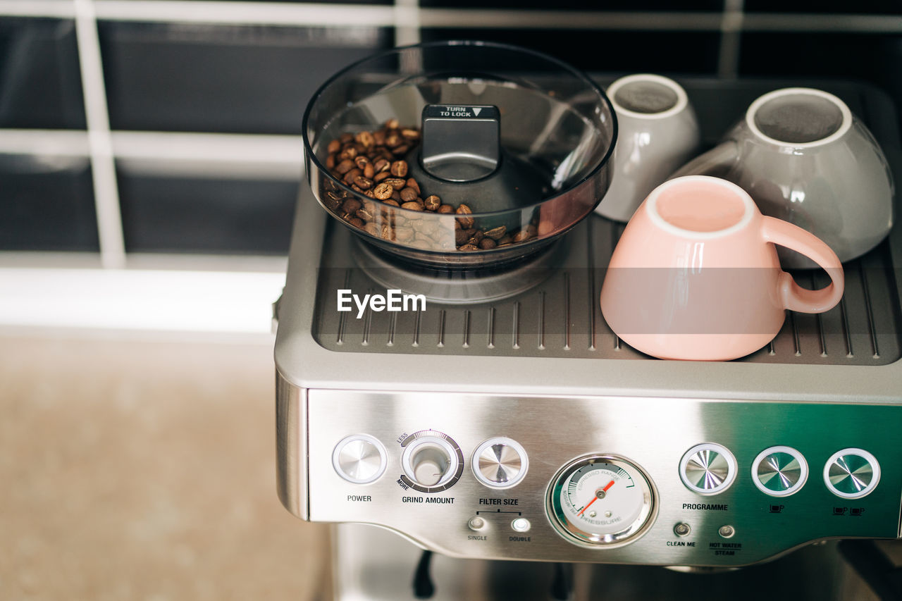 From above of roasted coffee beans in electric grinder against ceramic cups on rack of modern espresso maker at home