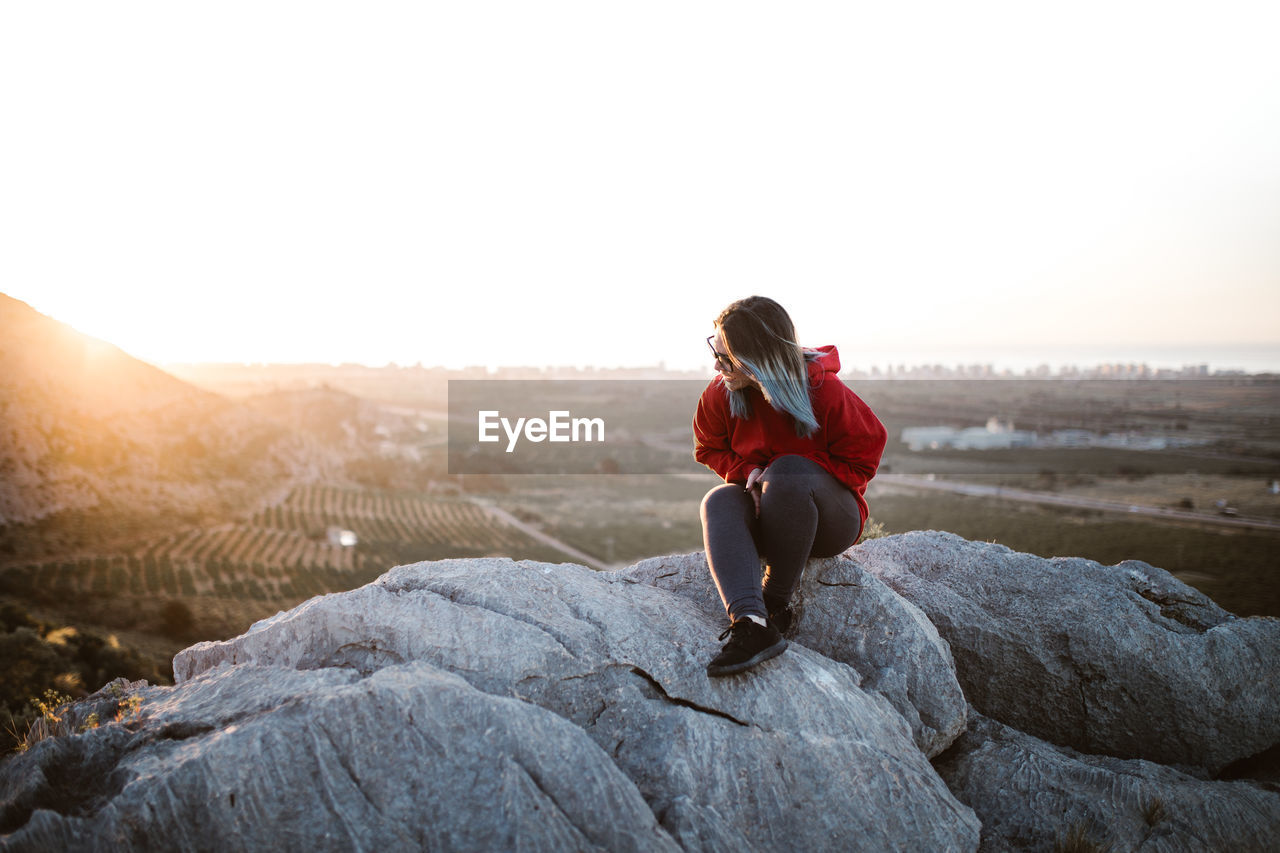 Picturesque view of young tourist sitting on peak of mountain and looking away in sunny day